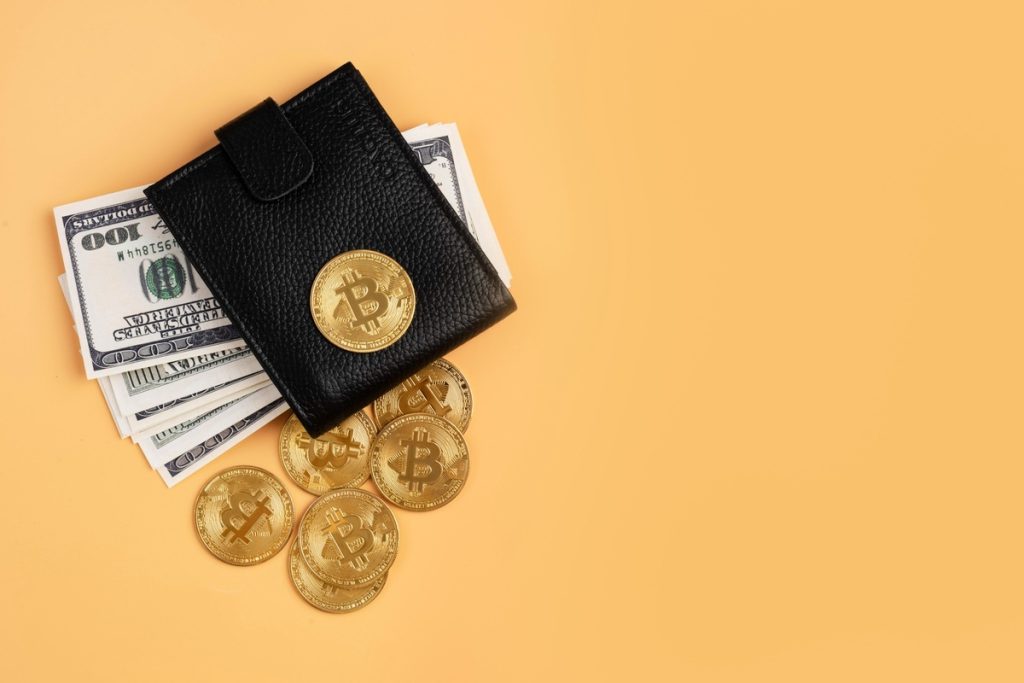 Black leather wallet placed over US dollar bills and several golden Bitcoin coins scattered on an orange surface. The composition evokes the concept of digital finance and cryptocurrency investment.