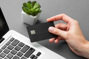 Hand holding a black credit card above the keyboard of a silver laptop with black keys, on a gray surface. In the background, there is a small green succulent plant in a white pot.