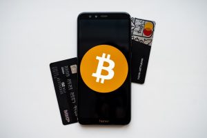 Black smartphone displaying the orange Bitcoin logo prominently on its screen, placed on a white surface. Behind the phone, there are two credit cards — one black and one gray — partially visible, suggesting the connection between cryptocurrencies and traditional payment methods.
